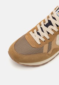 Barna suéd és hálós sneaker bézs fűzőkkel, gumitalppal és világosbarna díszítéssel. Texturált felsőrésze és ívelt orrformája van.