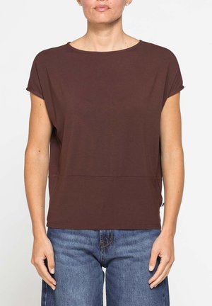 Persona che indossa una maglietta marrone a maniche corte semplice e jeans blu, in piedi davanti a uno sfondo bianco uniforme.