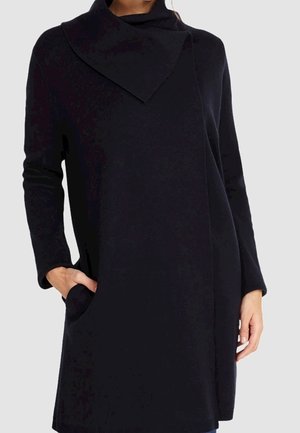 Femme portant un pull tunique noir à manches longues avec un col haut asymétrique et des poches latérales, une main dans la poche.