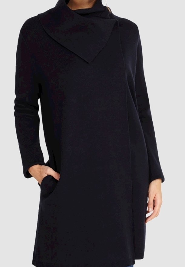 Femme portant un pull tunique noir à manches longues avec un col haut asymétrique et des poches latérales, une main dans la poche.