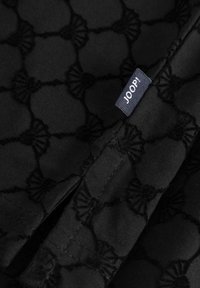 Tissu noir avec un motif en relief complexe ressemblant à des formes d'éventail, doté d'une étiquette bleue portant le texte "JOOP!" attachée.