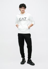 Mladý muž stojaci s rukami v kapsách, oblečený v bielej mikine EA7 Emporio Armani, čiernych teplákoch a čiernych teniskách na bielom pozadí.