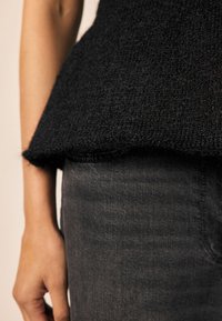 Fekete kötött pulóver, texturált felülettel, laza peplum alsó résszel. Sötét farmernadrágra van húzva. Az outfit közeli nézete.