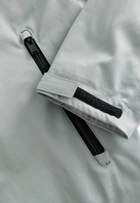 Materialul jachetei de un gri deschis are o suprafață texturată, având un fermoar negru și un accent din Velcro negru pe manșetă.