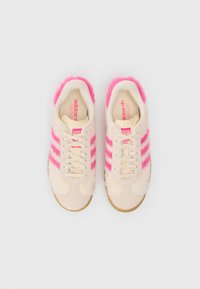 Lys beige sneakers med rosa pelskryss og stoffoverdel, med kremhvit snøring, polstrede såler og en teksturert gummisåle.