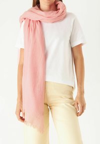 Écharpe rose clair en tissu doux, avec des bords à franges et un petit détail brodé de fleur. Portée sur une chemise blanche et un pantalon beige.