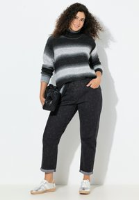 Šedý a černý pruhovaný svetr s vysokým límcem, v kombinaci s černými texturovanými kalhotami a stříbrnými teniskami, držící černou tašku.