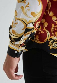 Manche d'une chemise à manches longues avec des motifs baroques rouges, noirs, blancs et dorés, portée par une personne en pantalon noir.