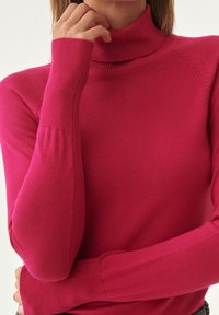 Dopasowany różowy sweter z golfem wykonany z miękkiego materiału, z prążkowanym kołnierzem i mankietami, o gładkiej fakturze i bezszwowym wykończeniu.