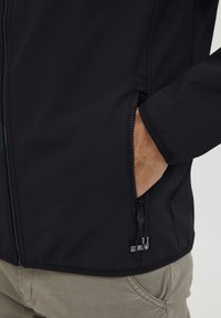 Crna softshell jakna s džepom na patentni zatvarač, s teksturiranim završetkom i izvezenim logotipom na donjoj strani.