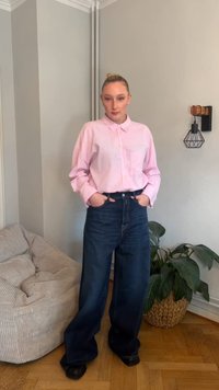 Camisa cor-de-rosa clara com botões e mangas compridas, enfiada em jeans azul-escuro de perna larga. Sapatos pretos e um sofá bege texturizado são visíveis.