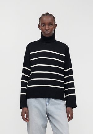 Femme portant un pull à col roulé noir avec des rayures horizontales blanches et un jean bleu clair, debout devant un fond clair uni.