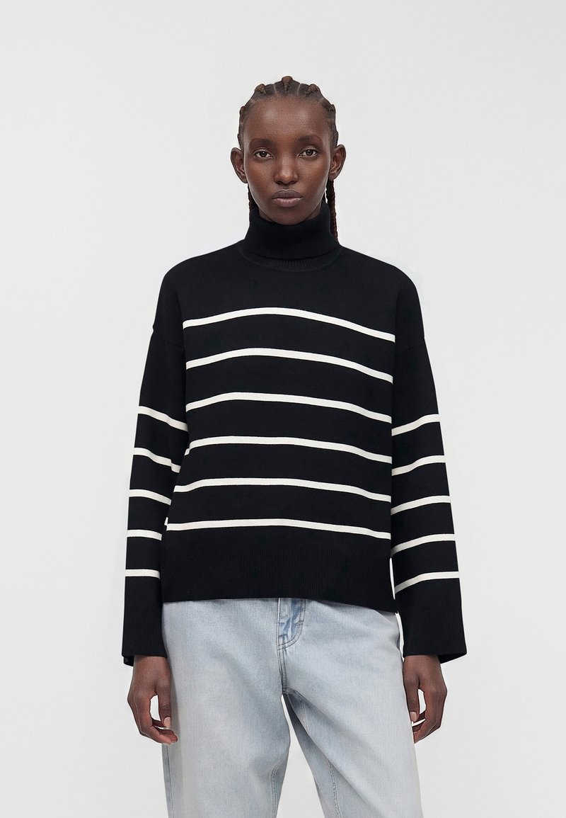 Donna che indossa un maglione nero a collo alto con righe orizzontali bianche e jeans azzurri, in piedi davanti a uno sfondo chiaro e uniforme.