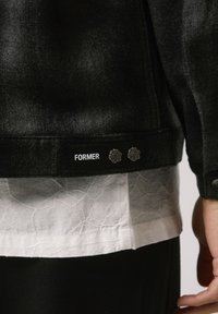 Veste noire avec une finition texturée, portant une chemise blanche superposée en dessous. Comprend une étiquette indiquant "FORMER" et deux accents métalliques en forme de rose.
