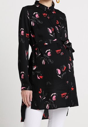 Mujer embarazada con una túnica negra de manga larga con un estampado floral colorido y pantalones blancos, de pie sobre un fondo liso.