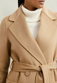 Abrigo de mezcla de lana color camel con cuello de muesca, cintura ceñida con cinturón y cierre de botones grandes. Presenta un acabado suave y texturizado.