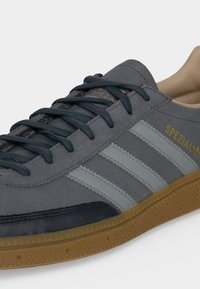 Γκρι σουέτ αθλητικό παπούτσι Adidas Spezial με σκούρα κορδόνια, λευκές ρίγες στο πλάι, κίτρινο λογότυπο και σόλα από καουτσούκ.