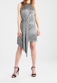 Robe sans manches argentée avec un devant drapé, un ourlet asymétrique et une texture douce ; associée à des talons hauts noirs à lanières.