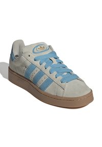 Hellgraue Wildledersneaker mit drei blauen Streifen, blauen Schnürsenkeln, einer runden Zehenpartie und einer strukturierten braunen Gummisohle. Goldene Logo-Akzent auf der Zunge.