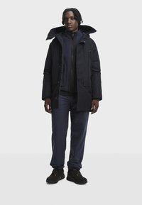 Man standing wearing black winter jacket, navy zip-up fleece, navy pants, and black hiking boots against a plain light background.