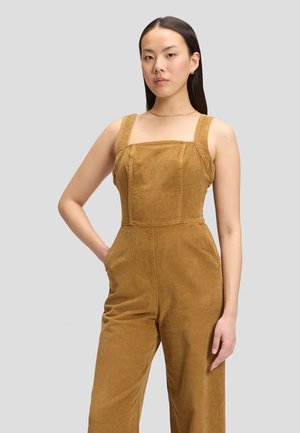 Femme aux longs cheveux foncés portant une combinaison sans manches en velours côtelé couleur camel avec jambes droites et poches, sur un fond clair uni.
