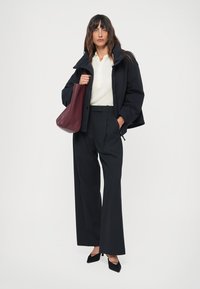 Vrouw staat in een witte top, zwarte pufferjack, ruime zwarte broek, met een grote bordeauxrode handtas in de hand en zwarte hakken.