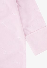 Roza flis jopica z teksturirano površino, ki vsebuje ponavljajoče se Nike logotipe. Rokavi imajo ravne kroje in čist zaključek.