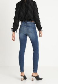 Jeans ajustados de mezclilla azul con cintura alta y cinco bolsillos, combinados con una blusa negra con flecos y mule negros de tacón con detalles en plateado.