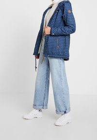 Blå mønstret jakke med hætte, der har et zigzag-design, fleece-for og snoretræk. Bæres med lyse denimjeans og hvide sneakers.