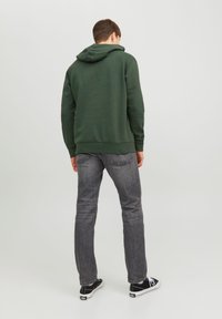 Felpa con cappuccio verde scuro e cordini, vestibilità rilassata, abbinata a jeans grigi e sneakers nere. Struttura semplice, texture liscia, design informale.