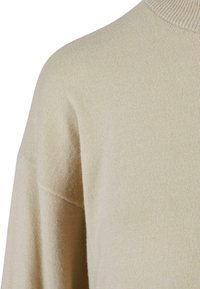 Beige neulepusero korkealla kauluksella, joka on pehmeää materiaalia, laskeutuvat hartiat ja väljä muotoilu.