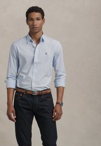 Svetlomodrá pruhovaná košeľa z bavlny s golierom na gombíky, v kombinácii s tmavými džínsami a hnedým koženým opaskom.