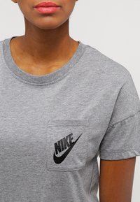 Šedé bavlněné tričko s kulatým výstřihem, krátkými rukávy a přední kapsou s černým potiskem loga Nike. Měkká textura.