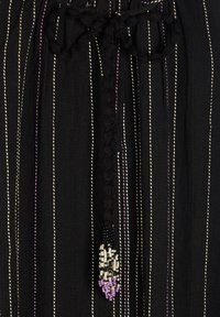 Czarny top z materiału z pionowymi paskami w białym i purpurowym kolorze. Zawiązywany na szyi z dekoracyjną frędzelką z koralików w kolorach czarnym, białym i purpurowym.