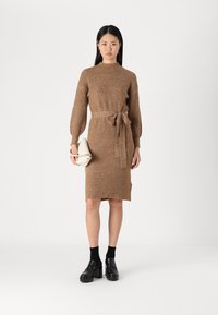 Vestito in maglia marrone, con collo alto, maniche lunghe, cintura in vita e texture a costine. Abbinato a scarpe con piattaforma nere.