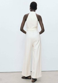 Une combinaison beige clair avec un col halter et des jambes larges, dotée d'une taille ceinturée et d'un tissu texturé. Portée avec des sandales plates.
