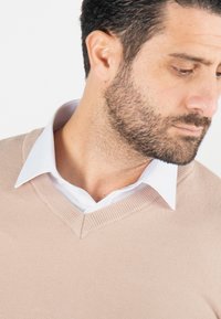 Pull en V beige superposé sur une chemise blanche. Le pull présente un ourlet et un col côtelés, une texture lisse et une coupe ajustée.