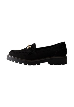 Mocasines de ante negro con suela gruesa y texturizada, y detalles en tono dorado. Presentan un diseño clásico y un ligero tacón elevado.