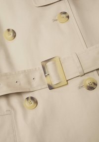 Beżowy materiał płaszcza trench z dużymi, okrągłymi, marmurkowymi guzikami oraz pasującym paskiem z prostokątną klamrą w stylu żółwia.