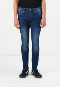 Vaqueros de mezclilla azul de corte slim con detalles descoloridos, diseño de cinco bolsillos y cierre de cremallera. Llevados con zapatillas negras y una camiseta negra.
