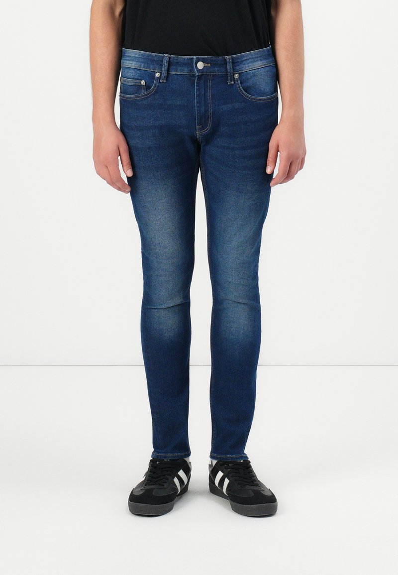 Vaqueros de mezclilla azul de corte slim con detalles descoloridos, diseño de cinco bolsillos y cierre de cremallera. Llevados con zapatillas negras y una camiseta negra.