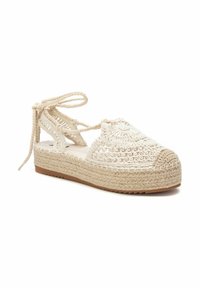 Sandalia de cuña de esparto en beige, con una parte superior de tejido trenzado, patrones de costura decorativos y lazos en el tobillo. Suela de yute natural.