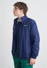 Tengerészkék színű széldzseki, cipzáros elülső résszel, könnyű anyagból, rugalmas mandzsettákkal és a mellrészen hímzett Adidas logóval.