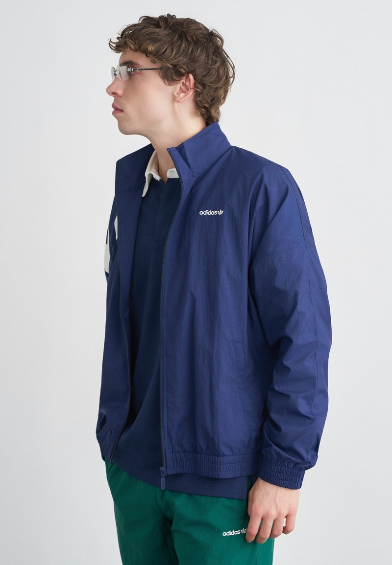 Tengerészkék színű széldzseki, cipzáros elülső résszel, könnyű anyagból, rugalmas mandzsettákkal és a mellrészen hímzett Adidas logóval.