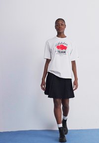 Hvid oversized t-shirt med rødt blomstermotiv og "Adidas" tekst, parret med en sort plisseret nederdel og sorte combat støvler.