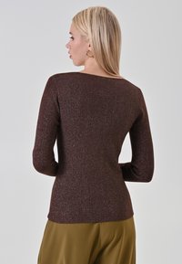 Femme blonde de profil portant des boucles d'oreilles en or, un pull à manches longues en maille marron, et un pantalon vert olive sur fond uni.