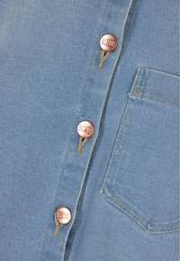 Tela de mezclilla azul clara con cuatro botones de presión de bronce a lo largo del tapacosturas y un bolsillo con costura simple en el lado izquierdo.