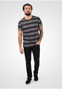 Svart stripete t-skjorte med hvite horisontale linjer, kombinert med sorte jeans og svarte joggesko, stående mot en ensfarget grå bakgrunn.