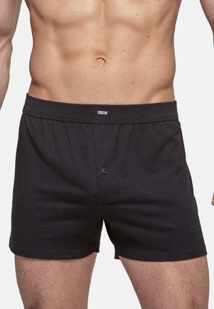 Mann trägt schlichte schwarze Knopf-Front-Boxershorts und zeigt Oberkörper und Oberschenkel vor einem weißen Hintergrund.