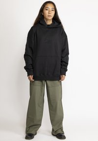 Zwarte oversized hoodie met een voorzak, voorzien van geborduurde details. Gepaard met olijfgroene wijde broekspijpen en zwarte schoeisel.